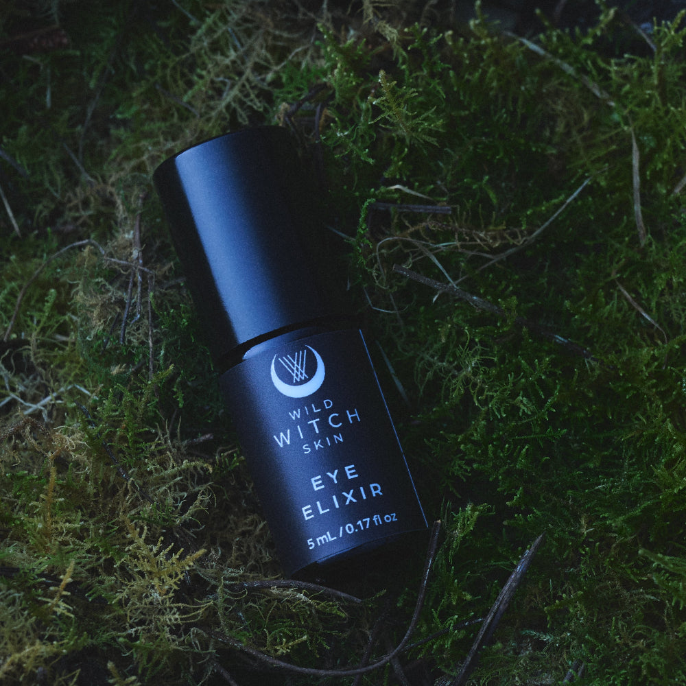 A black cylindrical bottle of Wild Witch Skin Eye Elixir lies diagonally on a bed of green moss and dried twigs.
