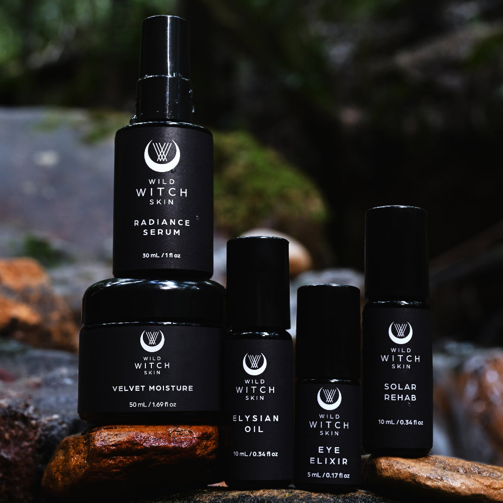Four black Wild Witch Skin cosmetic bottles and jars arranged on rocks in a forest setting. The products include Radiance Serum, Velvet Moisture,  Eye Elixir, Elysian Oil, and Solar Rehab.