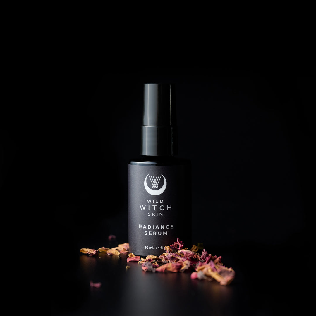 A black bottle of Wild Witch Skin Radiance Serum, styled with scattered dried pink and orange botanical petals on a reflective black surface against a solid black background.