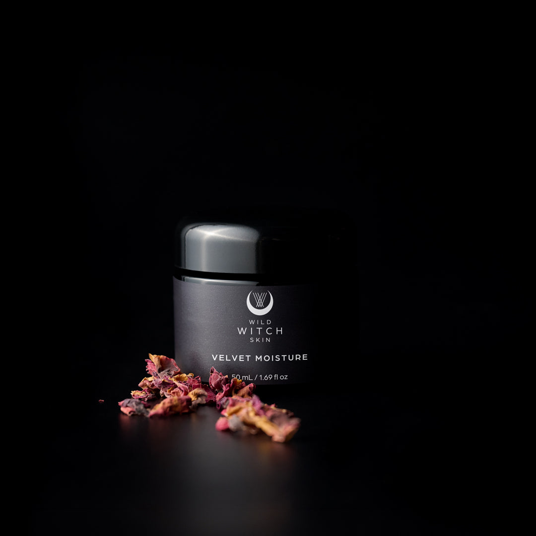 A black cosmetic jar labeled 'Wild Witch Skin Velvet Moisture' on a black background, with dried pink and orange flower petals placed in front of it.