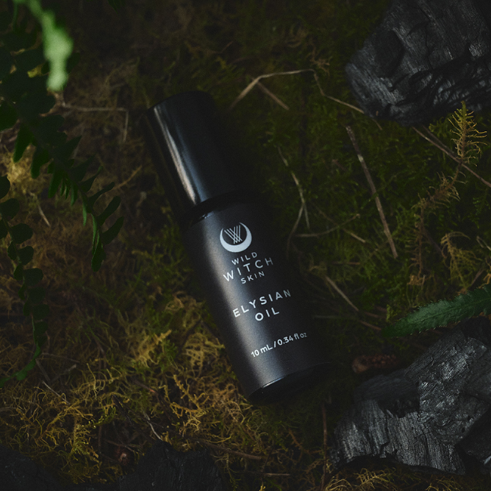 A black bottle of Elysian Oil by Wild Witch Skin lies on moss and dark rocks with green ferns, creating a moody, natural aesthetic.