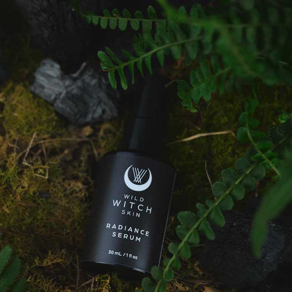 A black bottle of Wild Witch Skin Radiance Serum lies on mossy ground surrounded by green fern leaves and dark rocks.