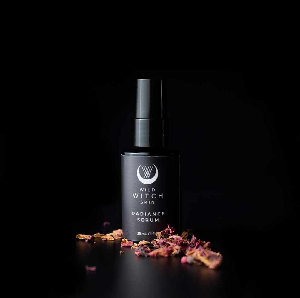 A black bottle of Wild Witch Skin Radiance Serum stands on a black surface with scattered dried pink and orange botanical pieces around its base, against a solid black background.