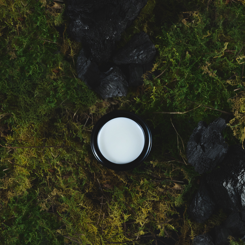 An open black cosmetic jar with a white cream inside sits on a bed of green moss and dark rocks, photographed from above.