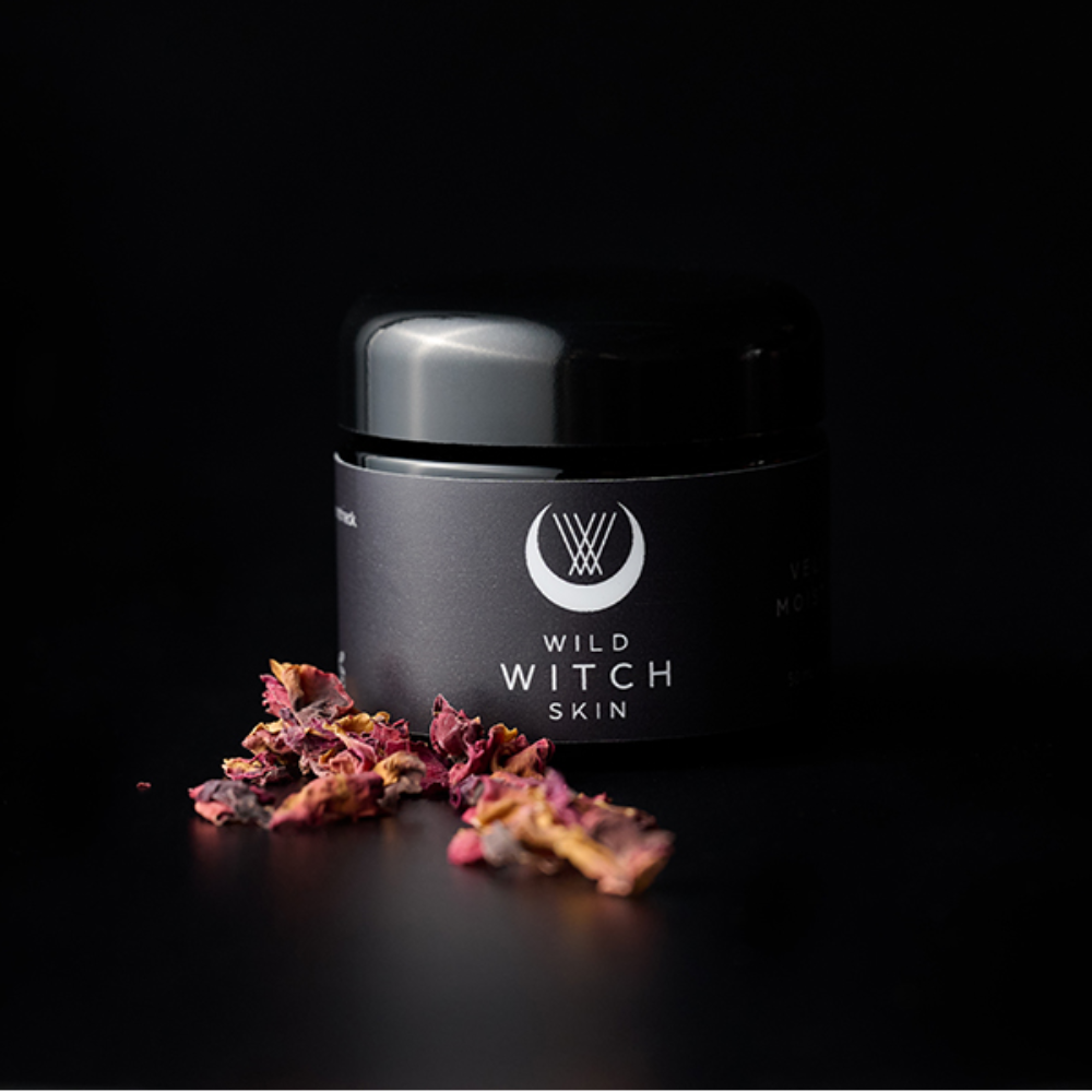 A black cosmetic jar labeled 'WILD WITCH SKIN' with a glossy lid, set on a black background with scattered dried rose petals in front.