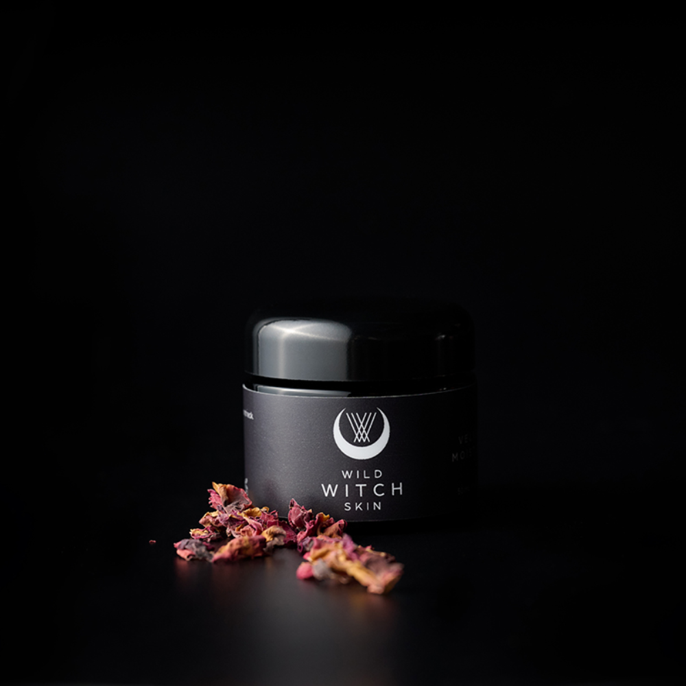 A black cosmetic jar labeled 'WILD WITCH SKIN' with a glossy lid, set on a black background with scattered dried pink and orange botanicals in front.