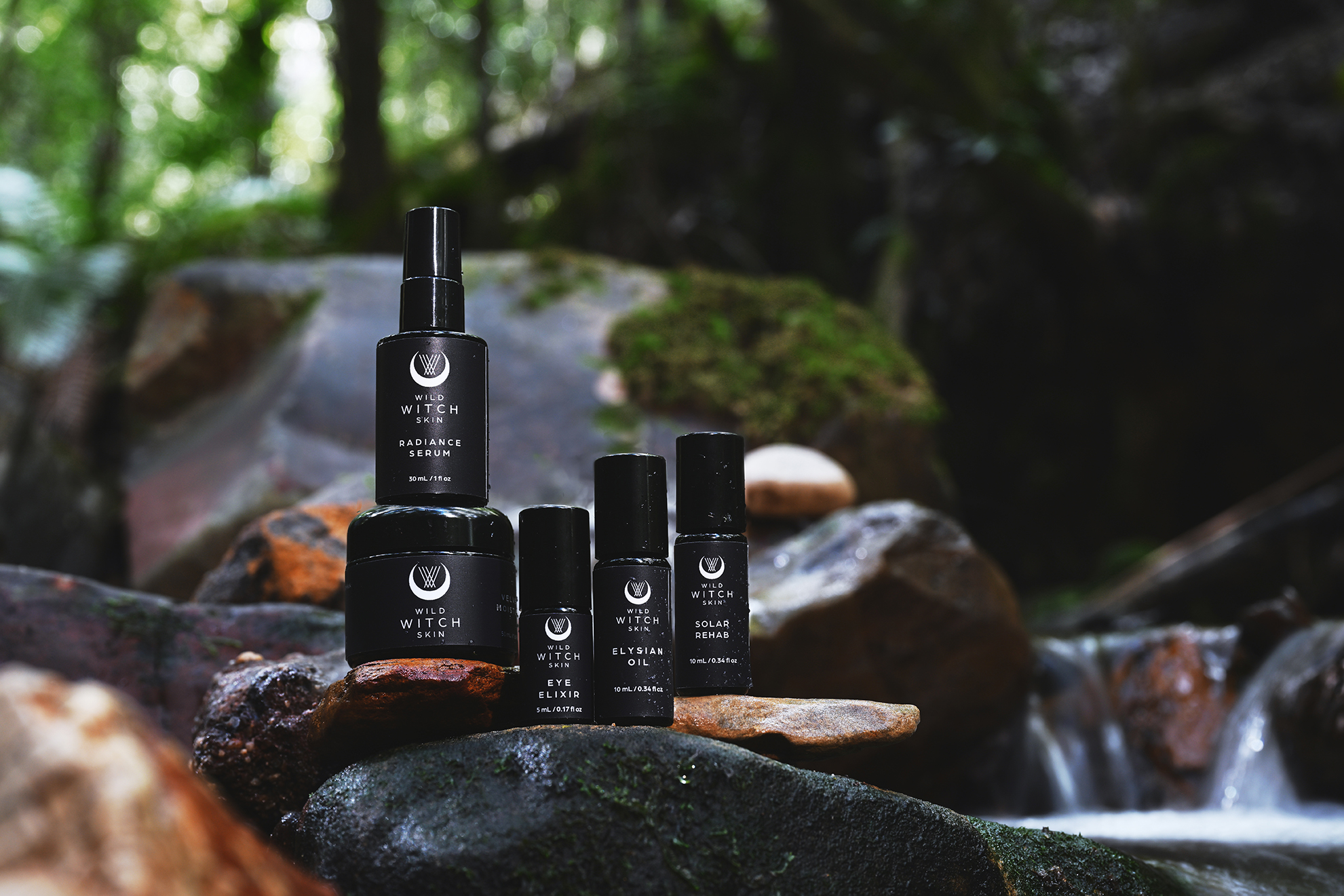 Five Wild Witch Skin black skincare bottles and jars arranged on mossy rocks beside a small forest stream.
