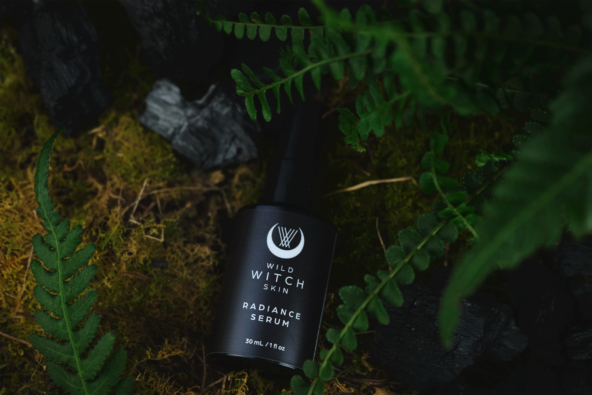 A black bottle of Wild Witch Skin Radiance Serum placed on mossy ground surrounded by green fern leaves and dark rocks.